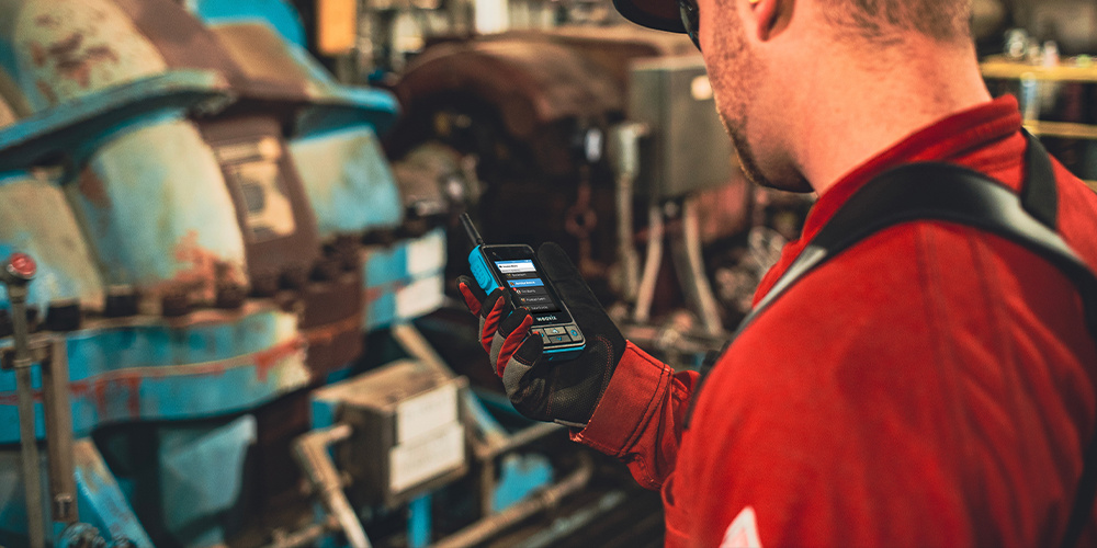 Thumbnail for a blog on how technology adoption empowers deskless workers. A worker wearing a hardhat, red shirt, and protective gloves looks at a rugged smart radio.